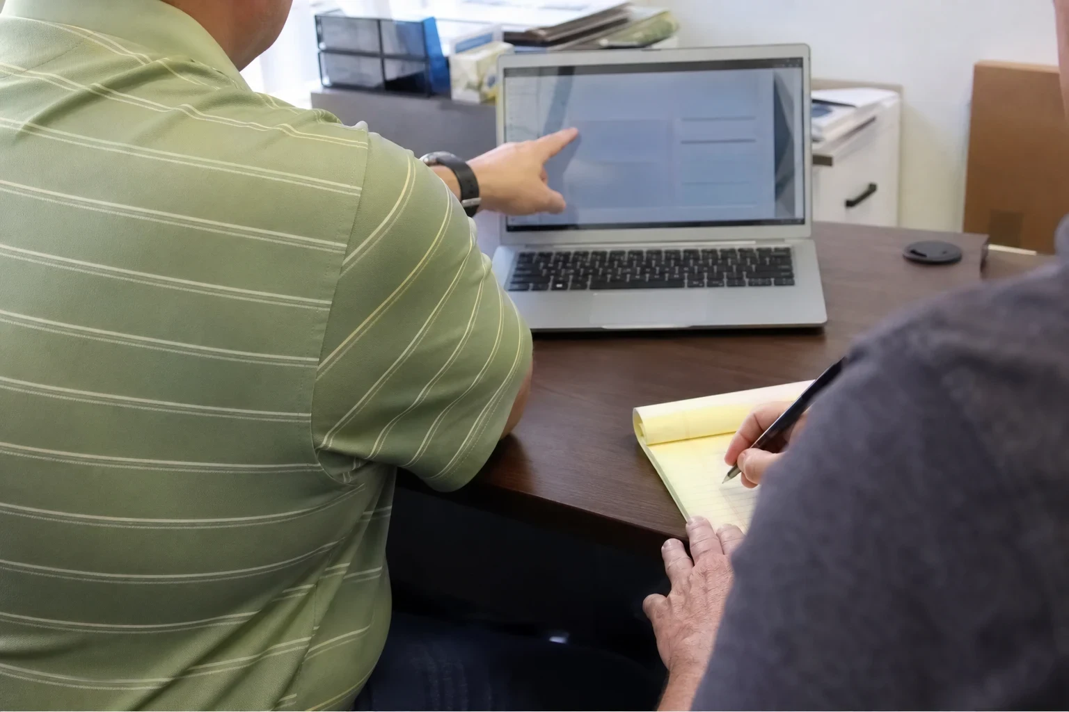 Over-the-shoulder view of a man in a green striped polo pointing at a laptop screen on an office desk while another man takes notes on a yellow legal pad.