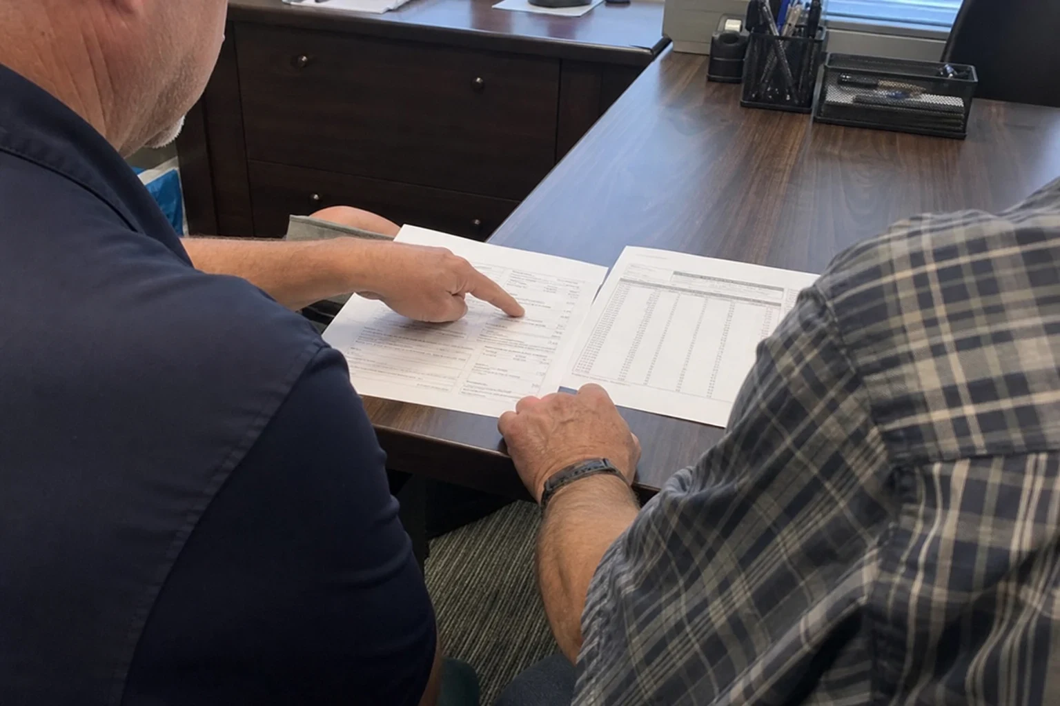 Two men seen from behind at a wooden office desk, one pointing at a statement laid beside a printed coverage table