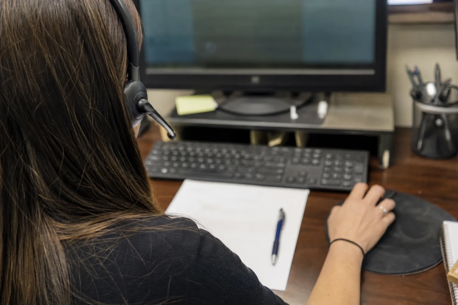 Over-the-shoulder view of an agent wearing a headset at a wooden desk, hand on a mouse, with a blank sheet of paper, a blue pen, keyboard and monitor